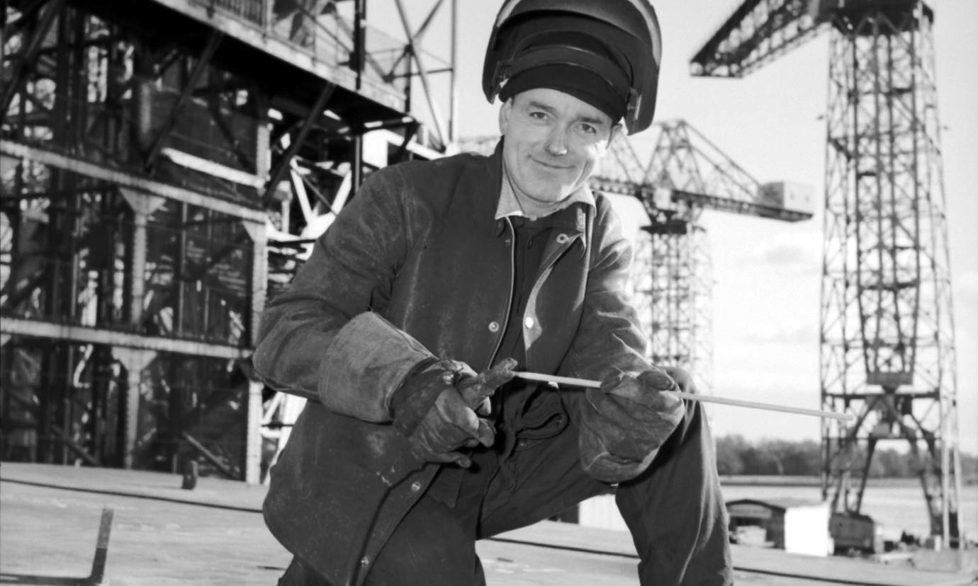 A shipyard welder, kneeling and wearing safety equipment and holding tools. In the background are cranes and scaffolding.