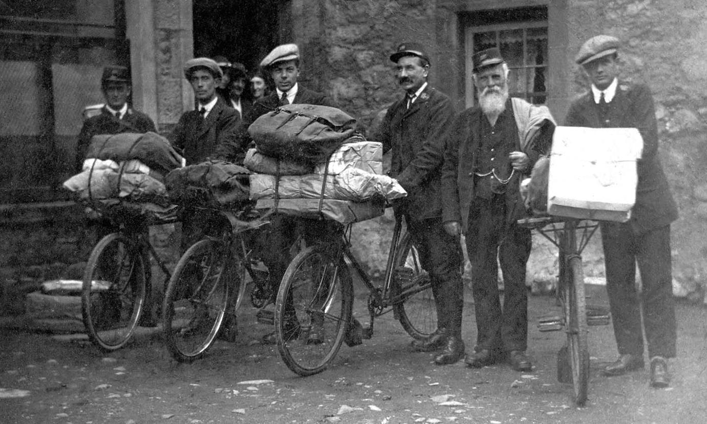 6 men are standing in smart clothes holding either sacks of post or bikes with mailbags and parcels.